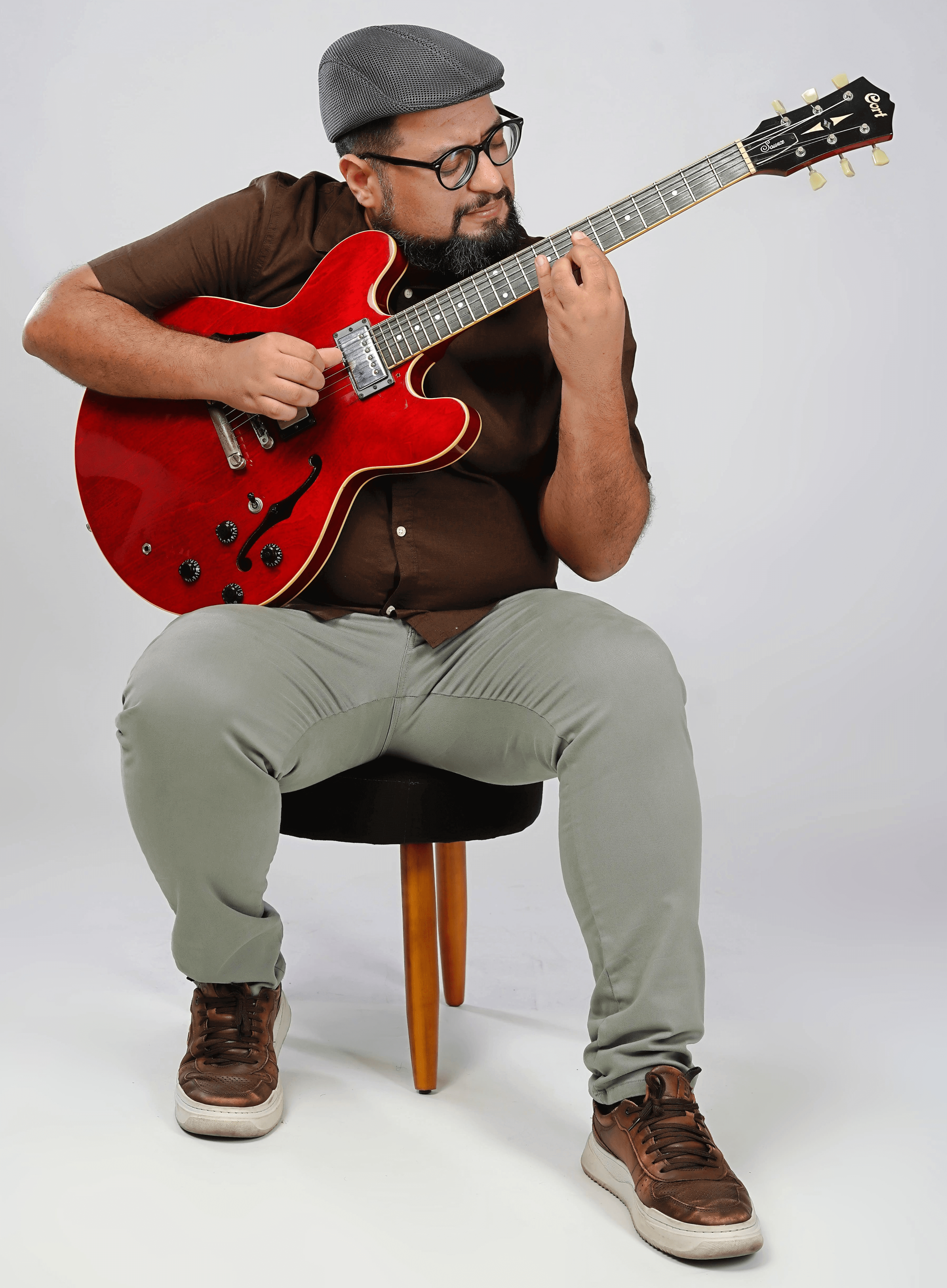 Liêve Ferreira tocando guitarra modelo ES-335 vermelha sentado em um banco contra um fundo branco